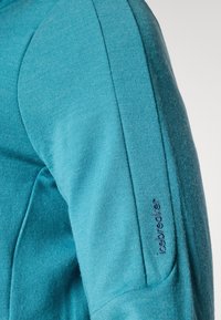 Teflonska majica z dolgimi rokavi v smaragdni barvi z teksturirano tkanino. Ima oblikovane ramene in zapleteno šivljenje po roki. Vidna blagovna znamka.