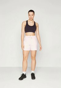 Frau steht da und trägt einen schwarzen Sport-BH, hellrosa Shorts mit Kordelzug, weiße Socken und schwarze Sportschuhe vor einem einfarbigen Hintergrund.