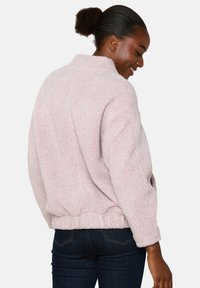 Rožinė fliso striukė su aukštu apykakle, elastinga juosmens juosta ir tekstūruotu audiniu. Išleista laisvu siluetu su ilgomis rankovėmis.