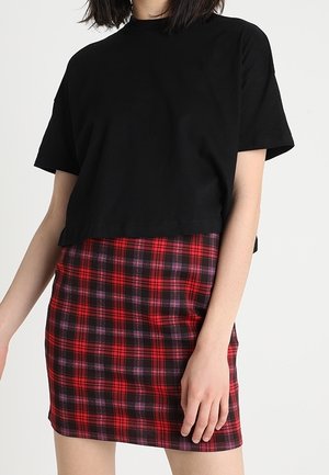 Kobieta ubrana w czarny krótki T-shirt i czerwoną, czarno kratowaną mini spódniczkę, stoi z lekko zgiętą ręką.