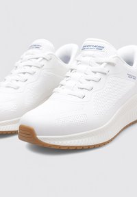 Białe sportowe buty z siateczkową cholewką, systemem sznurowania, teksturowanymi akcentami oraz gumową podeszwą. Logo znajduje się na języku.