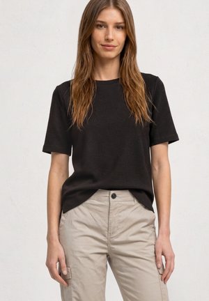 Junge Frau mit langen hellbraunen Haaren, die ein schwarzes strukturiertes Kurzarmshirt und beige Cargohosen trägt, steht vor einem einfarbigen Hintergrund.