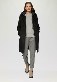 Zwarte lange jas met een capuchon, grijze gebreide sweater, grijze broek en tan enkellaarsjes. Glad materiaal en functionele ontwerpelementen.