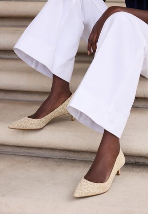 Personne portant un pantalon blanc à jambes larges et des chaussures beige à talons bas à motif, assise sur des marches en pierre.