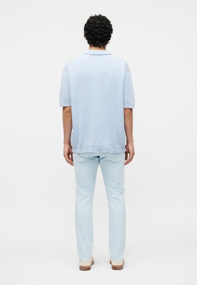 Άνδρας με σγουρά μαλλιά στέκεται με την πλάτη προς το μέρος μας, φοράει ανοιχτό μπλε κοντομάνικο πουκάμισο, ανοιχτό μπλε παντελόνι και μπεζ παπούτσια σε λευκό φόντο.