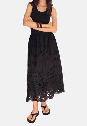 Femme portant un haut noir sans manches et une longue jupe noire avec un motif floral ajouré, debout les bras croisés et des sandales noires.