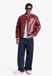 Bărbat purtând ochelari de soare negri, jachetă varsity din piele roșie, tricou alb, blugi largi bleumarin și adidași albi, stând cu o mână în buzunar.