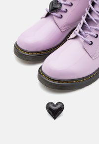 Fioletowe buty z patentowanej skóry z czarną gumową podeszwą, żółtymi szwami i fioletowymi sznurówkami. Mały czarny akcent w kształcie serca obok butów.