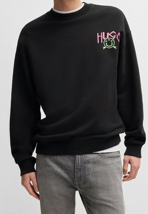 Zwarte trui met een geribbelde halslijn, met een felroze "HUGO"-logo en een groene smiley op de borst. Lange mouwen.