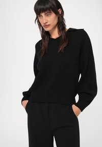 Fekete kötött pulóver V-nyakkal és leengedett vállakkal. A textúra bordázott, és laza fazonú, hosszú ujjú.