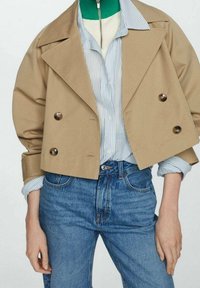 Cappotto trench cropped color cammello con chiusura a bottoni doppi, indossato sopra una camicia a righe blu chiaro, abbinato a jeans di denim blu.