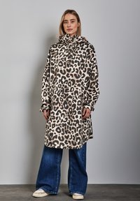 Abrigo impermeable de estampado de leopardo con cuello alto y capucha ajustable, combinado con jeans de pierna ancha en azul y zapatillas de color claro.