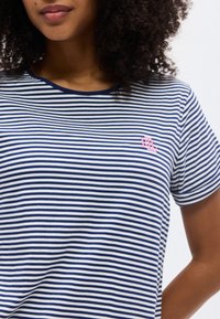 Γυναίκα με σγουρά μαλλιά που φοράει μπλε-ναυτικό και λευκό ριγέ T-shirt με ένα μικρό ροζ λογότυπο «RL» στο στήθος.