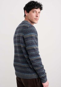 Vīrietis ar tumšiem, cirtainiem matiem, ģērbies rakstainā adītā džemperī, skatās pāri plecam pret neitrālu gaišu fonu.