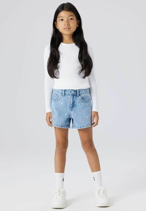 Ragazza con capelli lunghi e neri che indossa una maglia bianca a maniche lunghe, pantaloncini di jeans con stampe a forma di cuore, calzini bianchi e sneakers bianche, in piedi davanti a uno sfondo semplice.