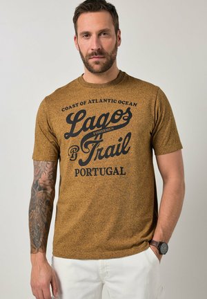 Mann mit Bart und tätowiertem Arm, der ein braunes gesprenkeltes T-Shirt mit schwarzem Text und weiße Hose trägt, Hand in der Tasche, neutraler Hintergrund.