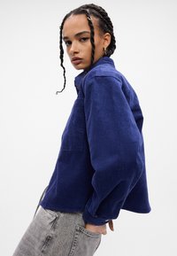 Camicia in corduroy blu navy con colletto e una tasca sul petto, dotata di maniche lunghe e una vestibilità rilassata. La texture è a coste e morbida.