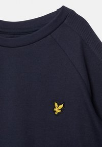 Marineblauer Strickpullover mit geripptem Rundhalsausschnitt und Schulterdetails, versehen mit einem kleinen gelben gestickten Logo eines fliegenden Adlers auf der Brust.