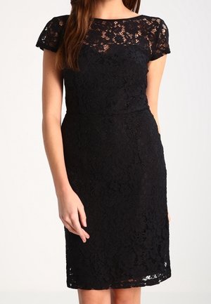 Vrouw draagt een nauwsluitende zwarte kanten jurk met korte mouwen en een doorschijnend kanten paneel op de bovenborst, staand tegen een effen lichte achtergrond.