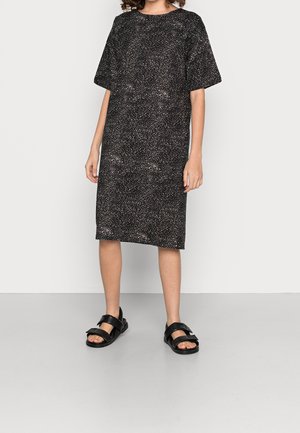 Młoda osoba ubrana w czarną sukienkę do kolan z białymi plamkami oraz czarne sandały z szerokimi paskami.