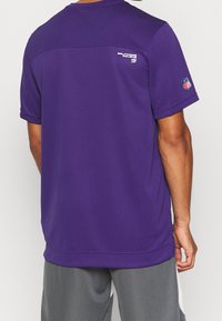 Fialové sportovní tričko s krátkým rukávem a kulatým výstřihem. Na zádech se nachází logo Baltimore Ravens a na levém rukávu je odznak NFL.