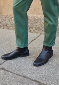 Bottes basses en cuir noir au design élégant, bout pointu et panneaux latéraux élastiques, portées avec un pantalon vert retroussé sur une surface en pierre.