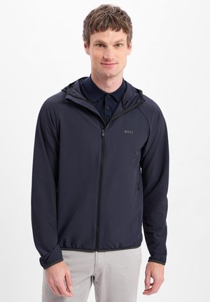 Man met kort bruin haar, gekleed in een donkerblauwe jas met rits over een donker overhemd met kraag en lichtgrijze broek, staand tegen een witte achtergrond.