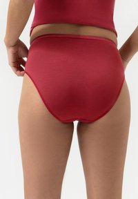 Persoon in een rode high-waist onderbroek en bijpassend topje, gezien vanaf de achterkant terwijl ze het kledingstuk aanpast tegen een witte achtergrond.