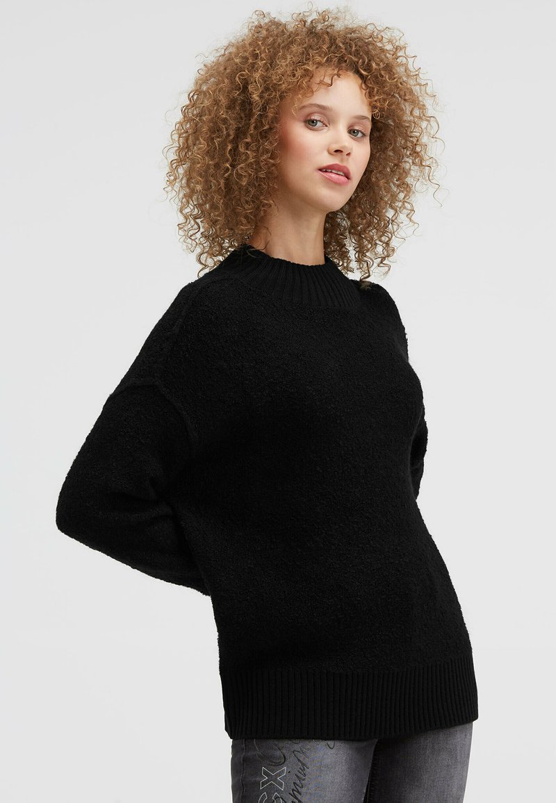 Frau mit lockigem Haar, die einen locker sitzenden schwarzen Pullover und graue Jeans trägt, vor einem schlichten hellen Hintergrund steht.