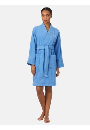 Donna in piedi a piedi nudi che indossa un accappatoio blu fino al ginocchio con cintura in vita e due tasche frontali su uno sfondo semplice.