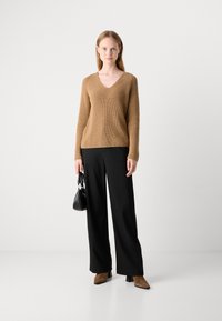 Pull en tricot côtelé marron avec un col en V, associé à un pantalon noir à larges jambes et des bottines en suède marron. La modèle tient un petit sac à main noir.