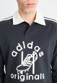Polo shirt negru cu mâneci lungi, cu guler alb și imprimeu grafic cu o încălțăminte. Prezintă trei dungi albe pe umeri.