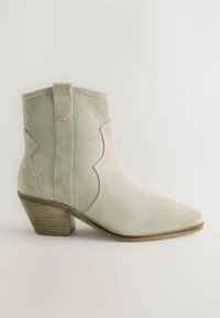 Botín de ante en gris claro con punta afilada, diseño lateral curvado y un tacón apilado texturizado. Superficie suave con un delicado bordado.