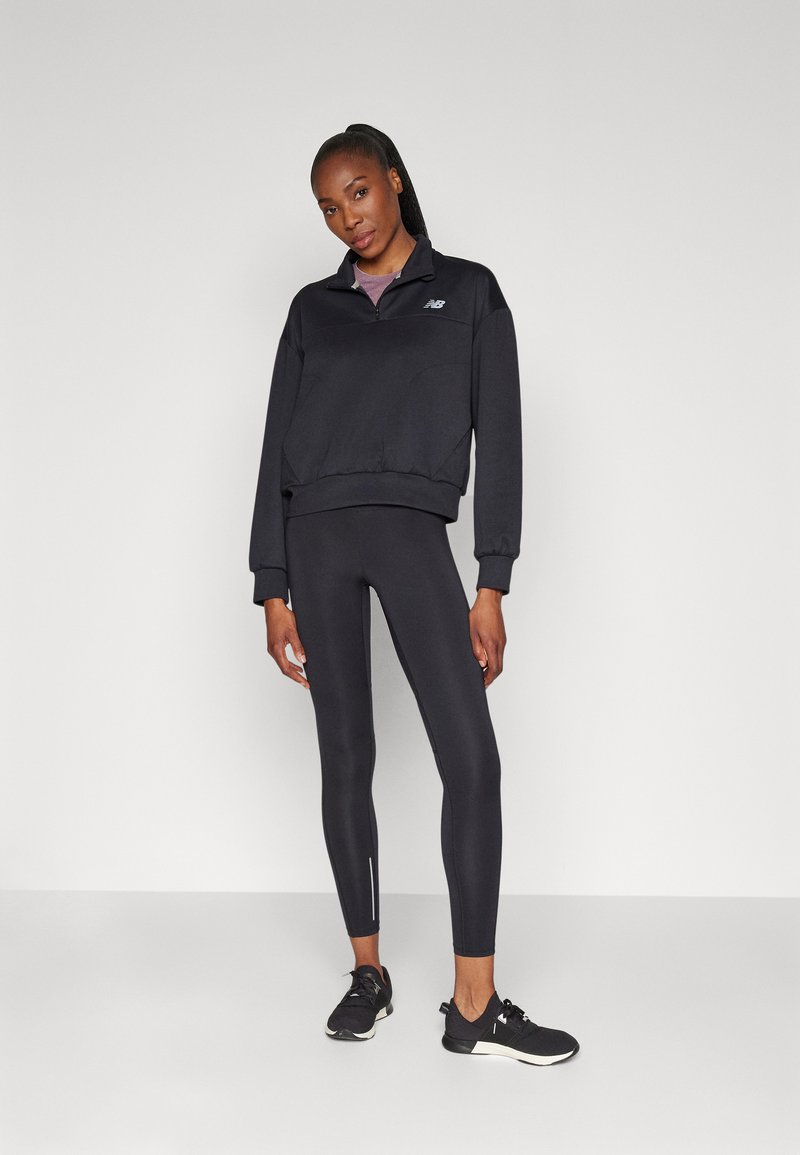 Felpa nera con zip e colletto alto, abbinata a leggings neri aderenti con un accento riflettente e scarpe da ginnastica nere.