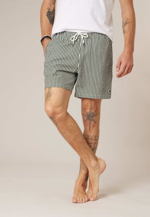 Homme portant un short rayé vertical noir et blanc avec cordon, debout pieds nus, montrant des tatouages sur les jambes et les bras.