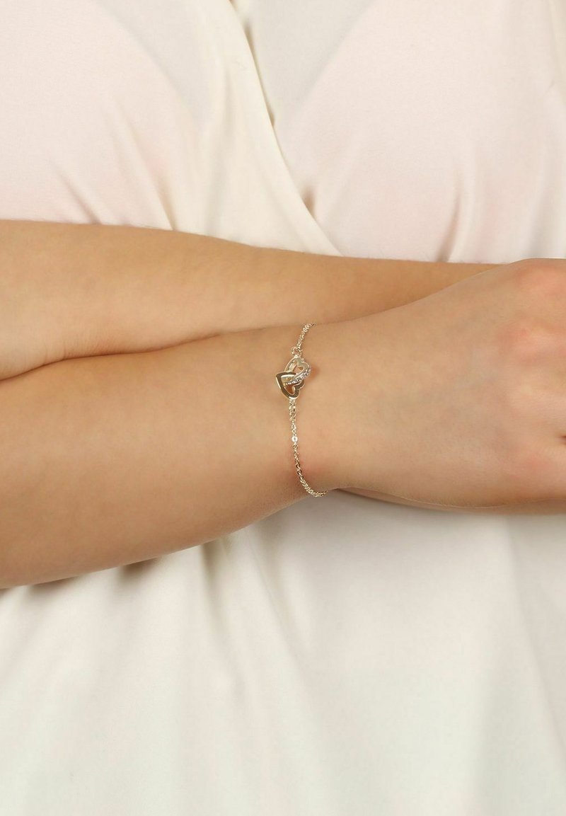 Silverkedja med ett hjärtformat hänge som har en klar sten, bärs på ett handled mot en ljus tygbakgrund.