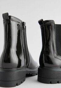 Botas de tobillo de charol negro con cremalleras laterales, paneles elásticos texturizados y suelas de goma gruesa con patrones de relieve profundos.
