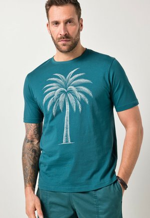 Mężczyzna o krótkich włosach i brodzie, ubrany w turkusowy T-shirt z dużym białym wzorem palmy i dopasowane turkusowe spodnie, stojący z jedną ręką w kieszeni.