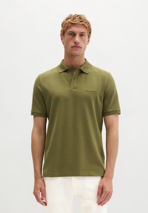 Mann trägt olivgrünes kurzärmeliges Poloshirt mit gesticktem Brustlogo, steht vor einfachem weißen Hintergrund.