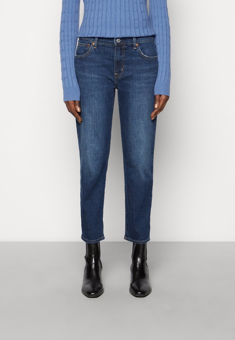 Personne portant un jean bleu taille mi-haute et coupe slim, un pull bleu clair en maille torsadée, et des bottines noires, debout sur un sol gris.