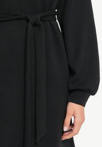 Vestido negro con un cinturón atado en la cintura, mangas voluminosas y una textura suave. Enfócate en el detalle del nudo del cinturón y los puños de las mangas.