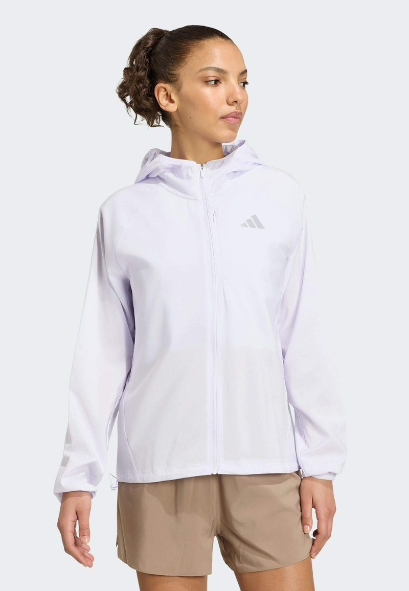Femme avec une queue de cheval bouclée portant une veste blanche à capuche zippée et un short beige, regardant légèrement vers sa droite.