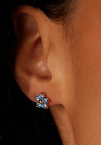 Gouden oorknop in de vorm van een bloem met zes lichtblauwe edelstenen en een gouden center, gedragen aan het oor. Close-up geeft de details van de zetting weer.