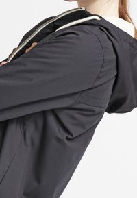 Kvinna som bär en svart huva jacka med en vit dragkedja, med armarna i kors och huvudet delvis synligt mot en vit bakgrund.