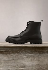 Bottine noire en cuir à lacets avec semelle en caoutchouc épaisse sur une surface en bois, fond uni.