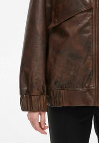 Pruun nahkjope koos tekstuuriga viimistluse, elastsete mansettide ja klapiga taskutega, osaliselt näidatud käe ja käega.