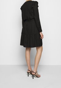 Schwarzes Kleid mit rüschiertem Ausschnitt und langen Ärmeln, ausgestattet mit einem elastischen Bund und einem gestuften Rock. Kombiniert mit schwarzen Riemchenschuhen.