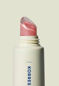 Bastone crema Korres con tubetto di plastica; il tappo trasparente a vite svela un prodotto rosa chiaramente scintillante. L'etichetta presenta testo blu su sfondo bianco.
