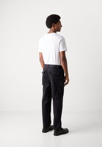 Pantalones cargo negros con bolsillos laterales, hechos de una tela duradera. Llevados con una camiseta blanca sencilla y zapatos negros, presentando un corte relajado.