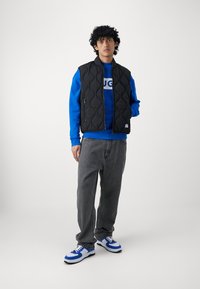 Schwarze, gesteppte Weste über einem blauen Sweatshirt, grauen Jeans und blau-weißen Sneakers. Die Weste hat einen Reißverschluss auf der Vorderseite und eine gesteppten Textur.